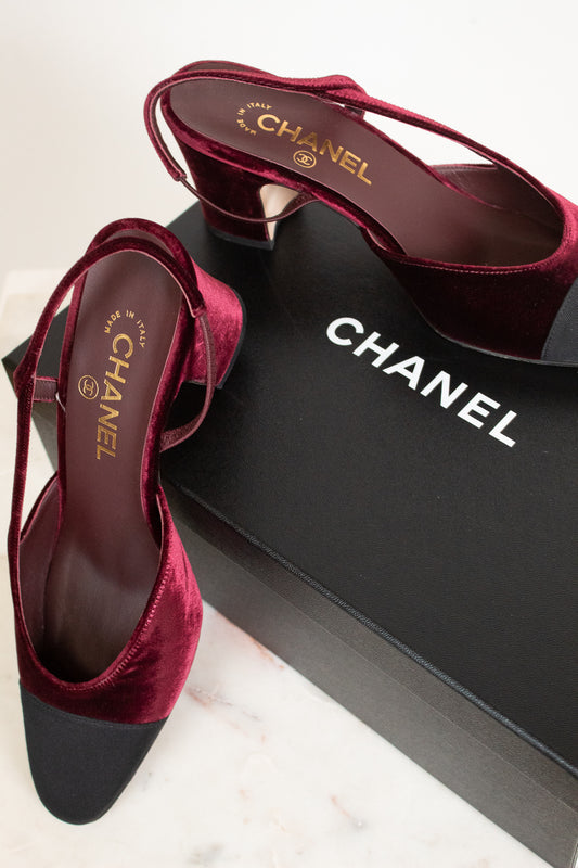 A pair of Chanel slingback high heels in deep burgundy velvet with black pointed toes, displayed on a black Chanel shoebox over a light marble surface.
