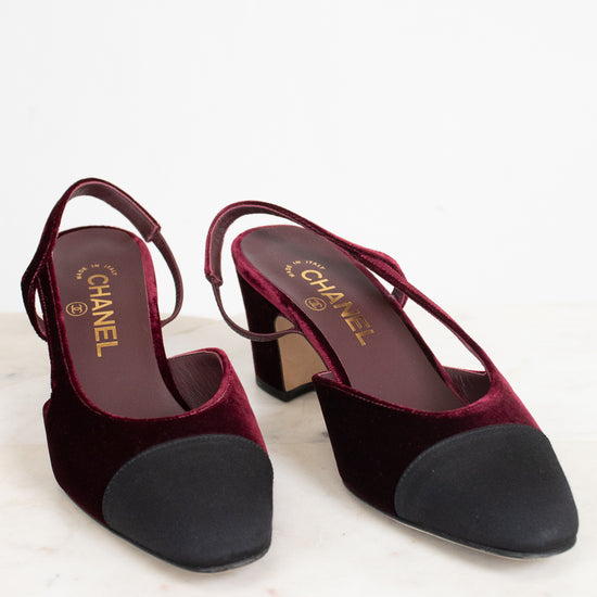 A pair of Chanel slingback block-heel pumps in deep burgundy velvet with black suede toe caps, displayed on a light marble surface against a white background.