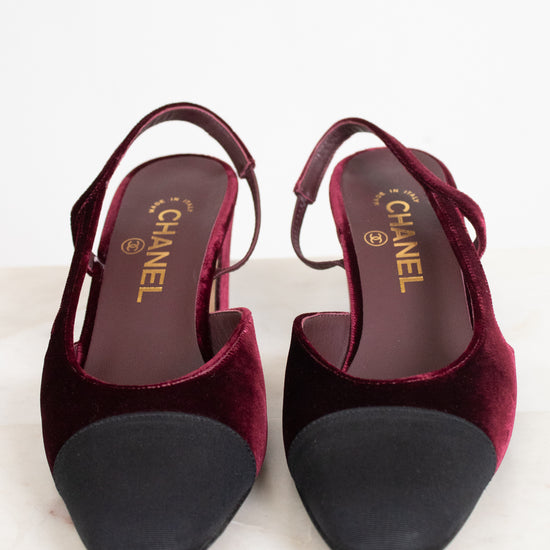 A pair of Chanel slingback pumps in deep burgundy velvet with black suede toe caps, shown from a top-down view on a light marble surface.