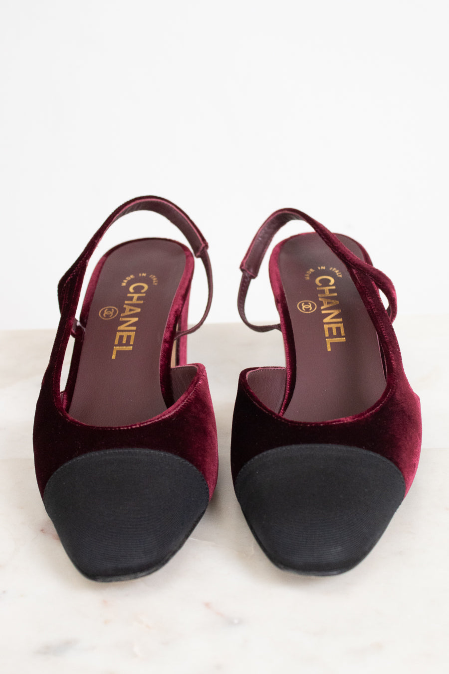 A pair of Chanel slingback pumps in deep burgundy velvet with black suede toe caps, shown from a top-down view on a light marble surface.