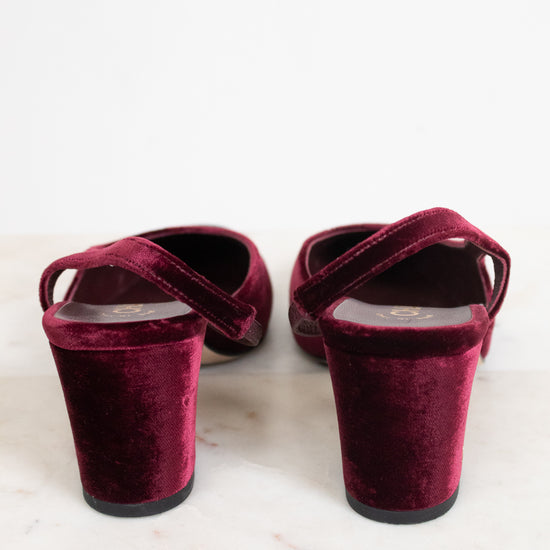 A pair of deep burgundy velvet slingback mule heels with block heels, shown from the back on a light marble surface against a white background.