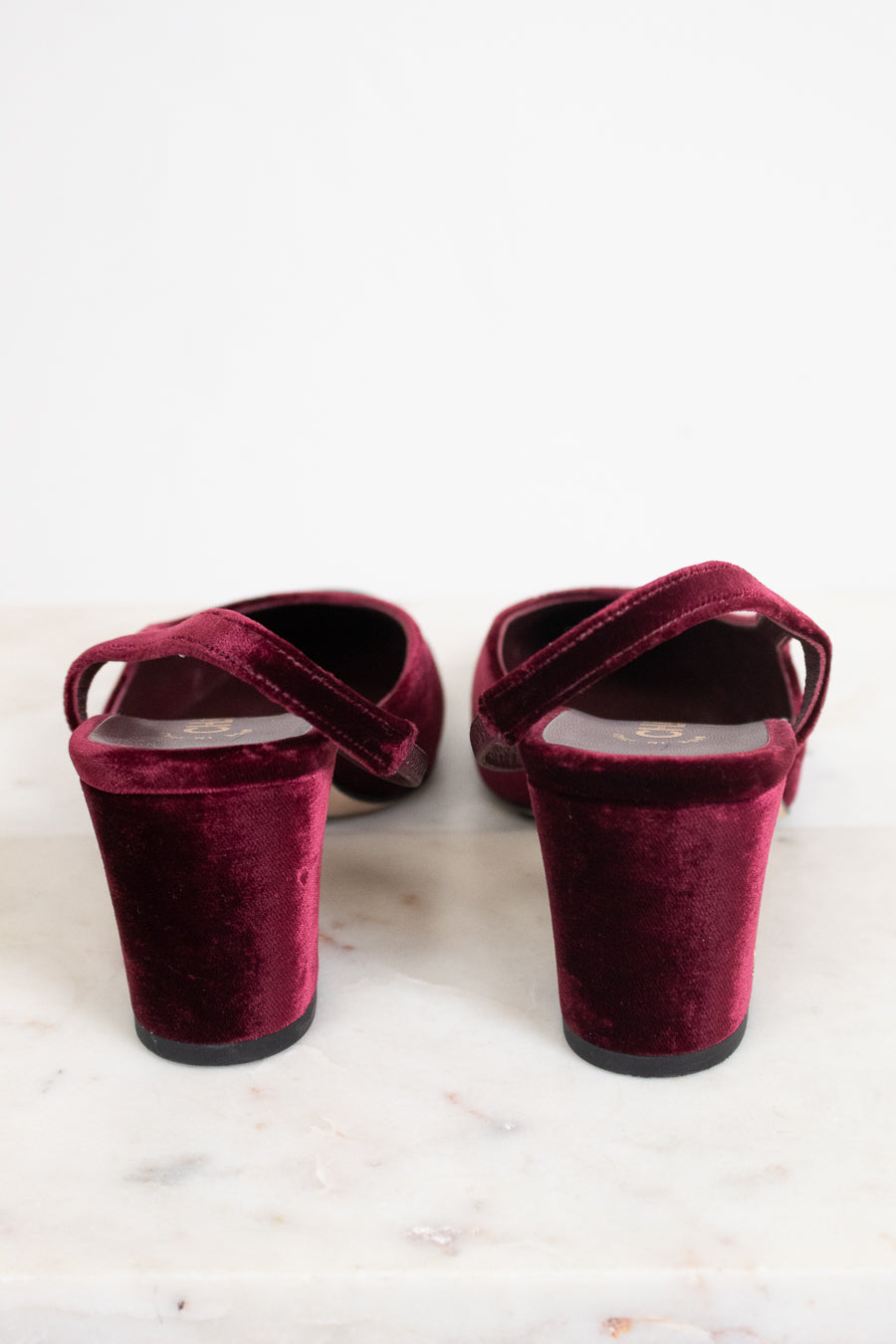 A pair of deep burgundy velvet slingback mule heels with block heels, shown from the back on a light marble surface against a white background.