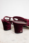 A pair of Chanel slingback block-heel sandals in deep burgundy velvet, featuring a gold-tone interlocking CC logo on the heel and a gold insole logo, displayed on a light marble surface against a white background.