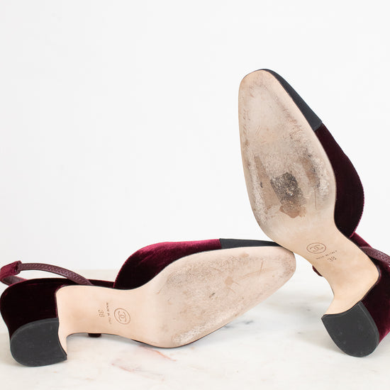 A pair of women's slingback heels shown from the sole, revealing a deep burgundy velvet upper, a beige insole, and black rubber soles, resting on a white marble surface against a white background.