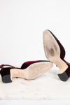A pair of women's slingback heels shown from the sole, revealing a deep burgundy velvet upper, a beige insole, and black rubber soles, resting on a white marble surface against a white background.