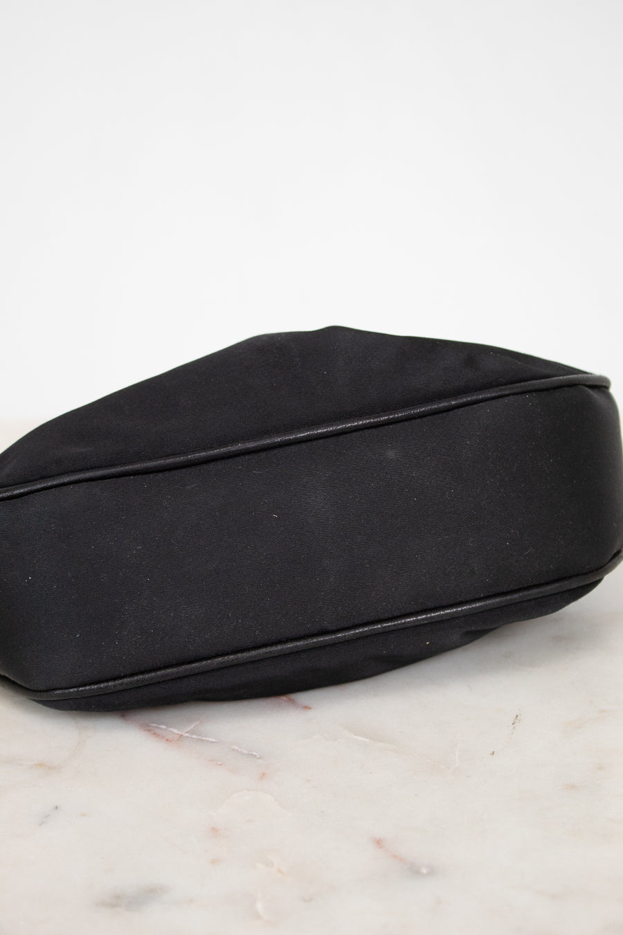 A black satin pouch bag with black leather piping, resting on a white marble surface against a white background.