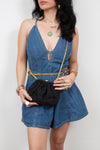 A woman wearing a blue denim romper with a keyhole cutout and gold chain details, holding a small black pouch bag with a gold chain strap.
