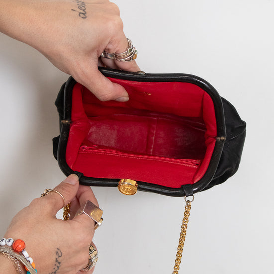 A black structured clutch handbag with a bright red interior lining and a gold-tone chain strap, shown open with two hands holding it against a plain light background.