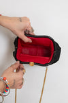A black structured clutch handbag with a bright red interior lining and a gold-tone chain strap, shown open with two hands holding it against a plain light background.
