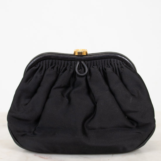 A black gathered fabric clutch bag with a gold-tone metal clasp, displayed on a light marble surface against a white background.