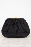 A black gathered fabric clutch bag with a gold-tone metal clasp, displayed on a light marble surface against a white background.