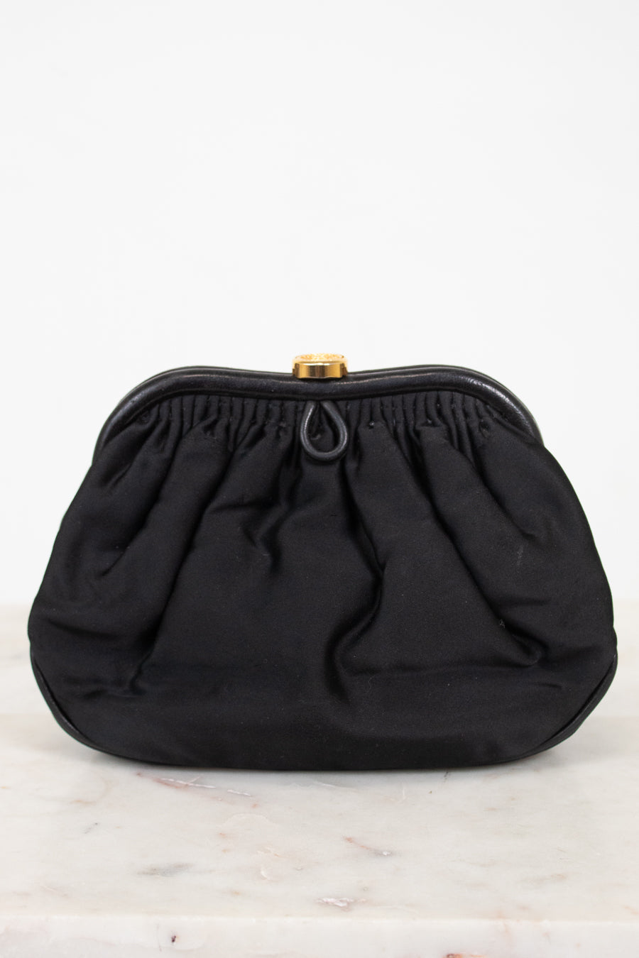 A black gathered fabric clutch bag with a gold-tone metal clasp, displayed on a light marble surface against a white background.