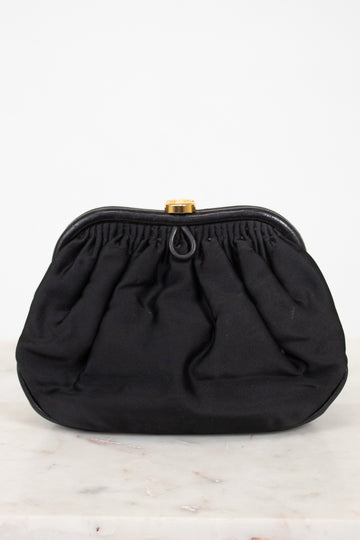 A black gathered fabric clutch bag with a gold-tone metal clasp, displayed on a light marble surface against a white background.