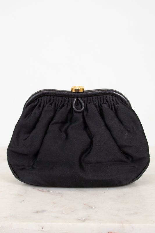 A black gathered fabric clutch bag with a gold-tone metal clasp, displayed on a light marble surface against a white background.