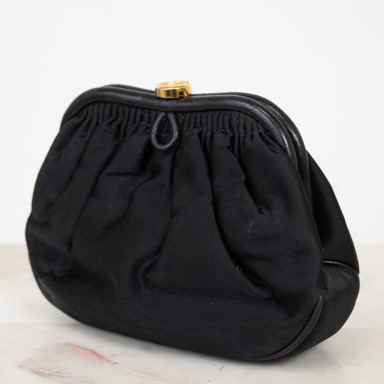 A black fabric clutch purse with a gathered, pleated design and a small gold-tone metal clasp, resting on a light marble surface against a white background.