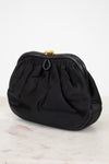 A black fabric clutch purse with a gathered, pleated design and a small gold-tone metal clasp, resting on a light marble surface against a white background.