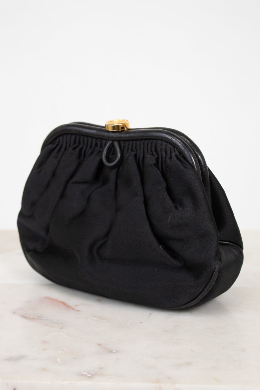 A black fabric clutch purse with a gathered, pleated design and a small gold-tone metal clasp, resting on a light marble surface against a white background.