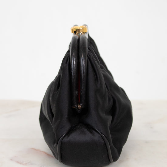 A black gathered clutch bag with glossy dark piping and gold-tone hardware, photographed from the side on a light marble surface against a white background.