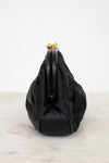 A black gathered clutch bag with glossy dark piping and gold-tone hardware, photographed from the side on a light marble surface against a white background.