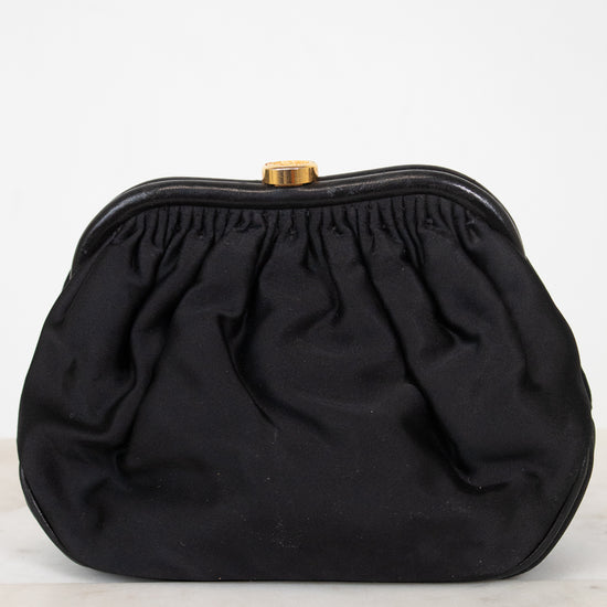 A black gathered clutch bag with a gold-tone metal clasp, resting on a light marble surface against a white background.