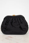 A black gathered clutch bag with a gold-tone metal clasp, resting on a light marble surface against a white background.