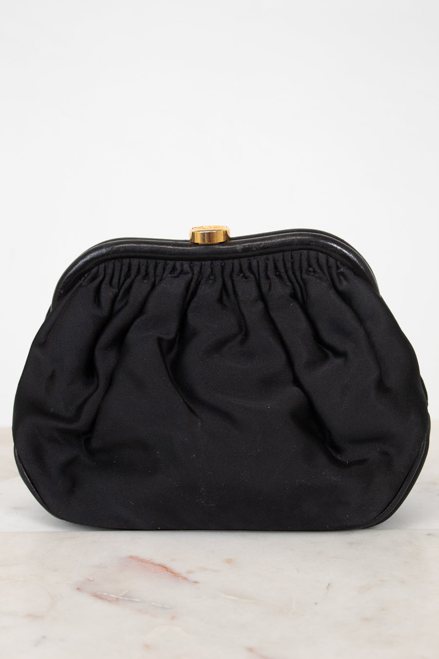 A black gathered clutch bag with a gold-tone metal clasp, resting on a light marble surface against a white background.