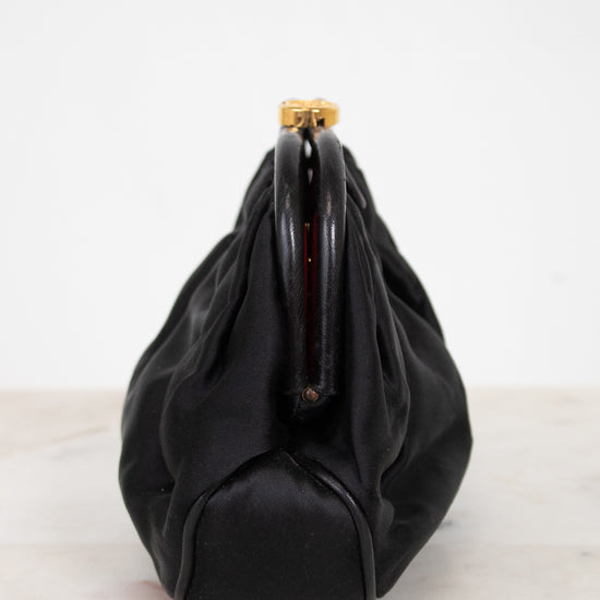 A black leather and satin shoulder bag with a gathered, slouchy silhouette and a gold-tone metal clasp closure, shown from the side on a light marble surface against a white background.