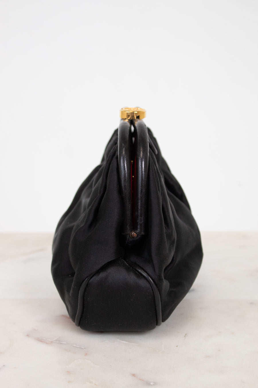A black leather and satin shoulder bag with a gathered, slouchy silhouette and a gold-tone metal clasp closure, shown from the side on a light marble surface against a white background.