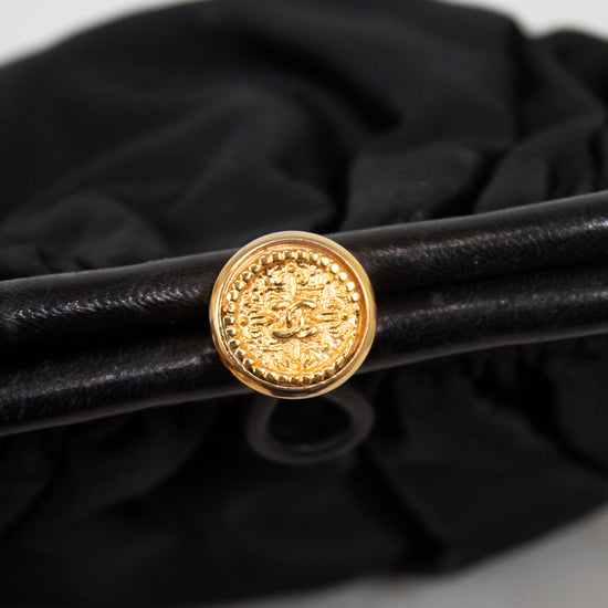 A close-up of a black leather bag with a prominent round gold-tone decorative clasp, set against a black fabric background.