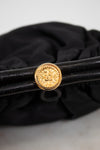 A close-up of a black leather bag with a prominent round gold-tone decorative clasp, set against a black fabric background.