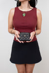 Person wearing a burgundy top and black skirt holding a black Chanel wallet against a plain background