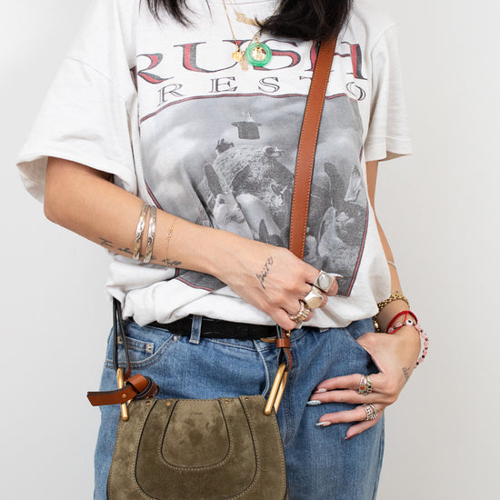Person wearing a graphic t-shirt and holding an olive leather bag against a plain background