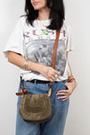 Person wearing a graphic t-shirt and holding an olive leather bag against a plain background