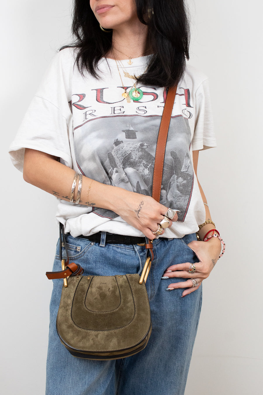 Person wearing a graphic t-shirt and holding an olive leather bag against a plain background