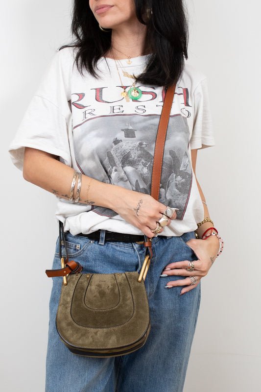 Person wearing a graphic t-shirt and holding an olive leather bag against a plain background