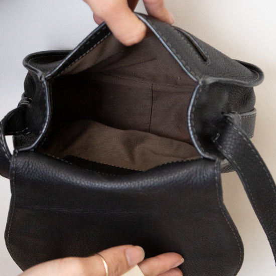 A close-up of a black pebbled leather bag being opened by two hands, showing the dark brown interior lining. The person wearing gold rings holds the bag open against a plain white background.
