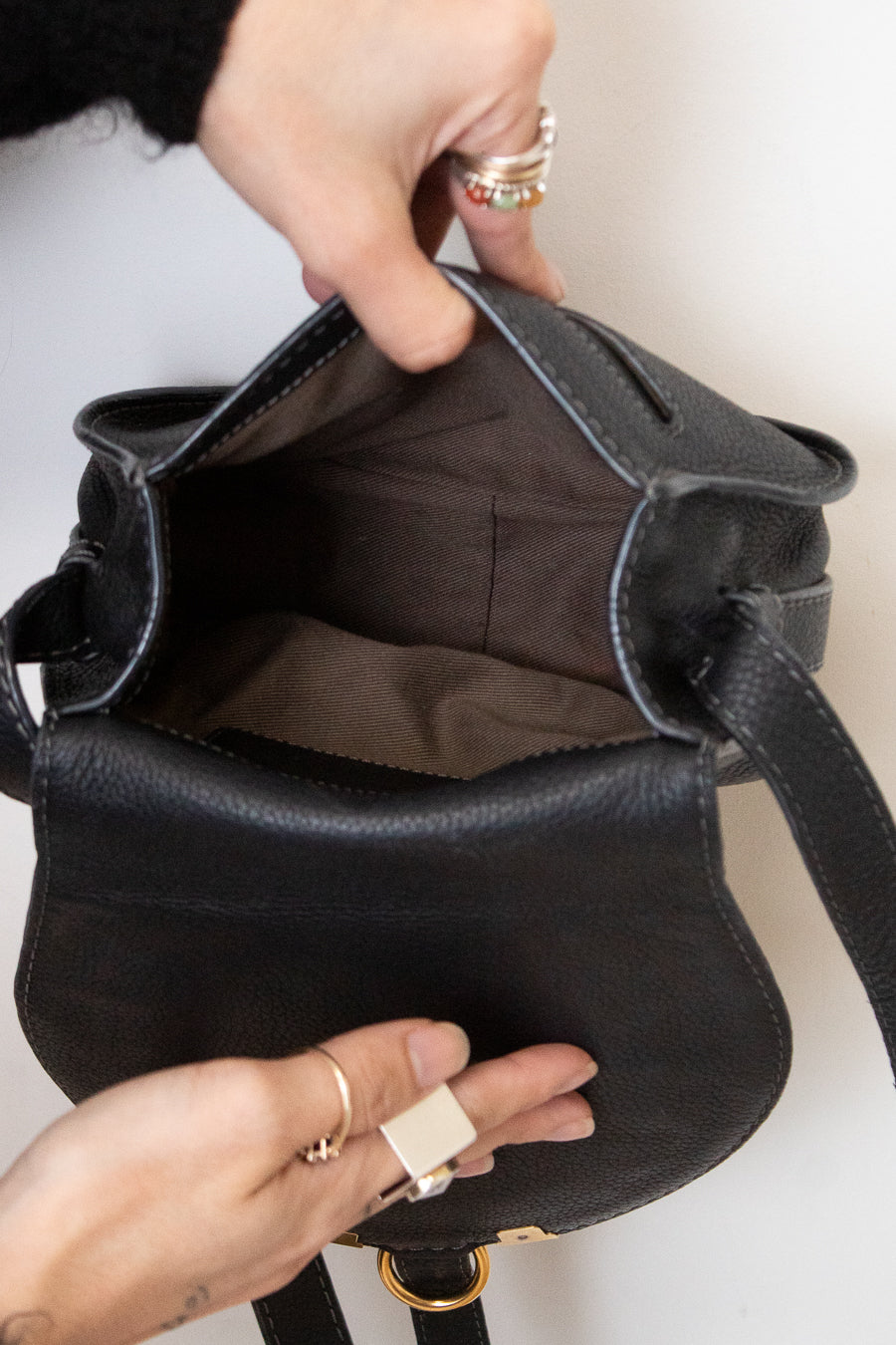 A close-up of a black pebbled leather bag being opened by two hands, showing the dark brown interior lining. The person wearing gold rings holds the bag open against a plain white background.