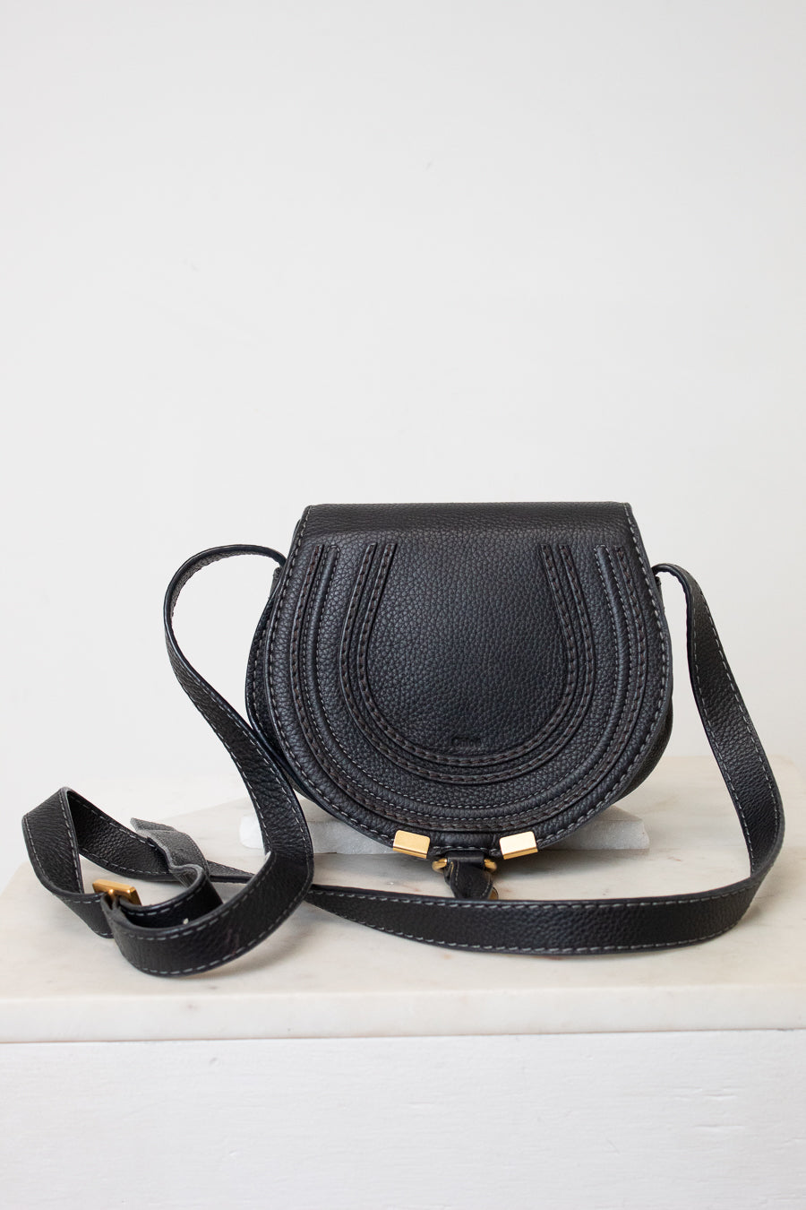A black pebbled leather crossbody bag with a curved, saddle-like silhouette, featuring gold-tone hardware and a matching black strap, displayed on a white surface against a white background.