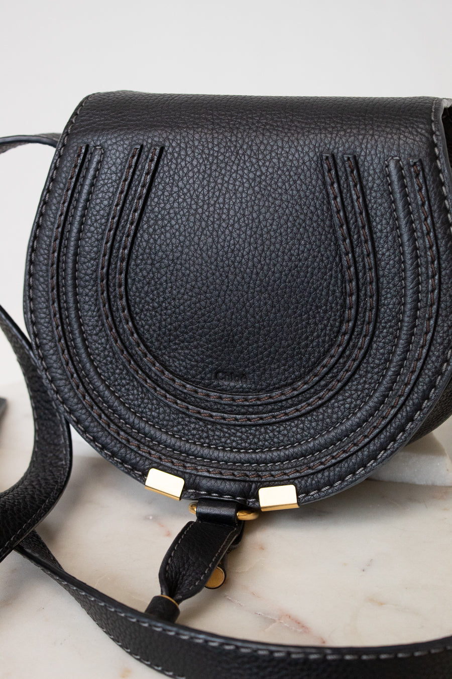 A black pebbled leather saddle bag with gold-tone hardware, resting on a light marble surface.