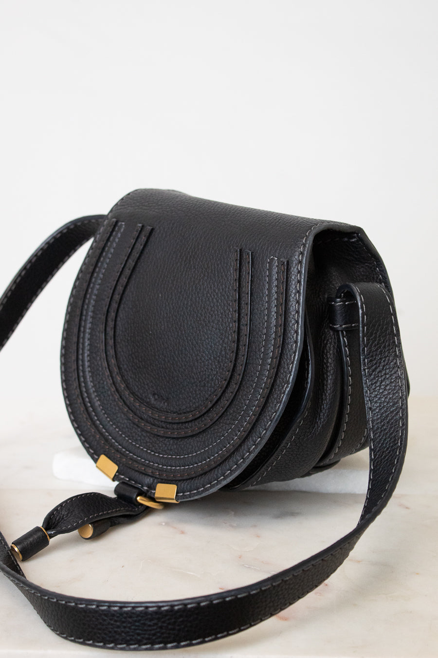 A black pebbled leather saddle bag with gold-tone hardware and a matching black leather shoulder strap, resting on a light marble surface against a white background.