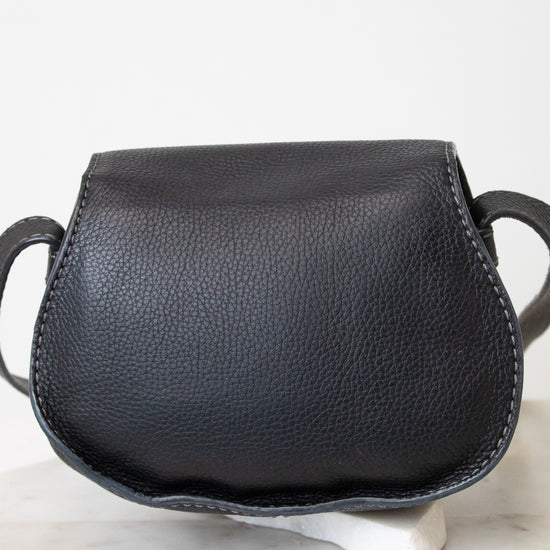A small black pebbled leather shoulder bag with a curved, saddle-like silhouette, displayed on a white ceramic stand over a light marble surface against a white background.