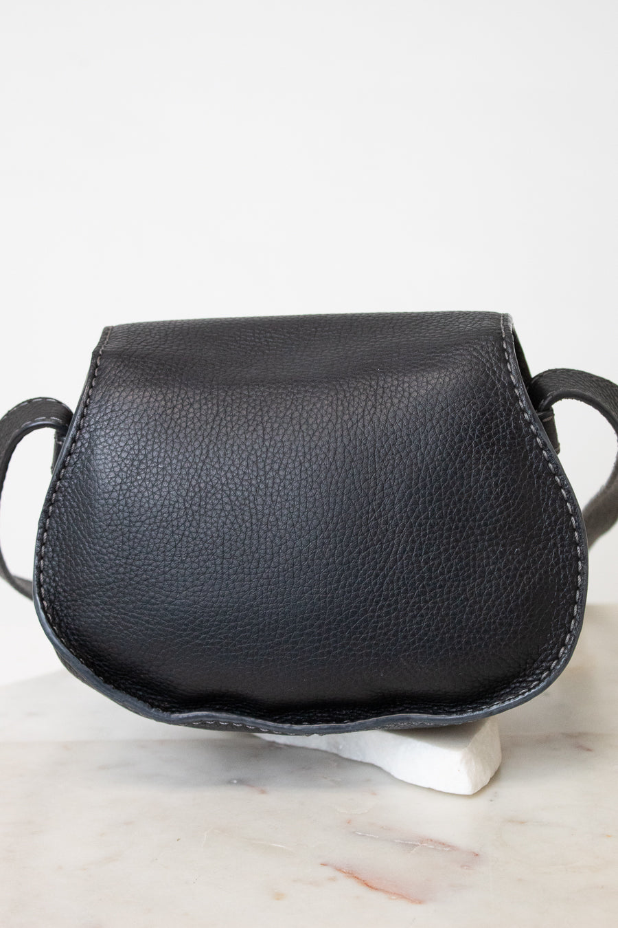 A small black pebbled leather shoulder bag with a curved, saddle-like silhouette, displayed on a white ceramic stand over a light marble surface against a white background.