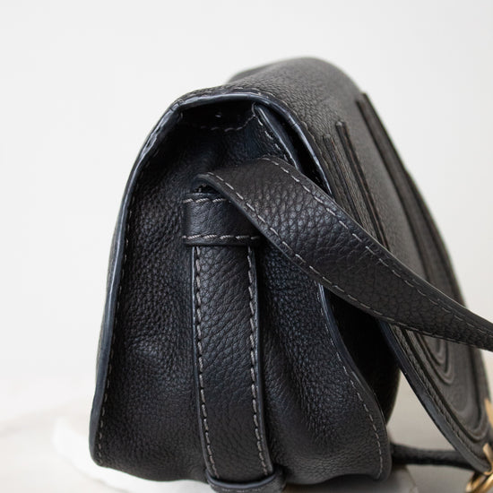 A close-up side view of a black pebbled leather handbag resting on a white marble display block against a clean white background.