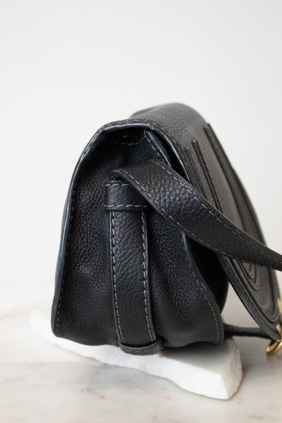 A close-up side view of a black pebbled leather handbag resting on a white marble display block against a clean white background.