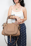 A woman wearing a white camisole and navy polka-dot pants holds a tan leather handbag with gold-tone hardware against a plain light gray background.