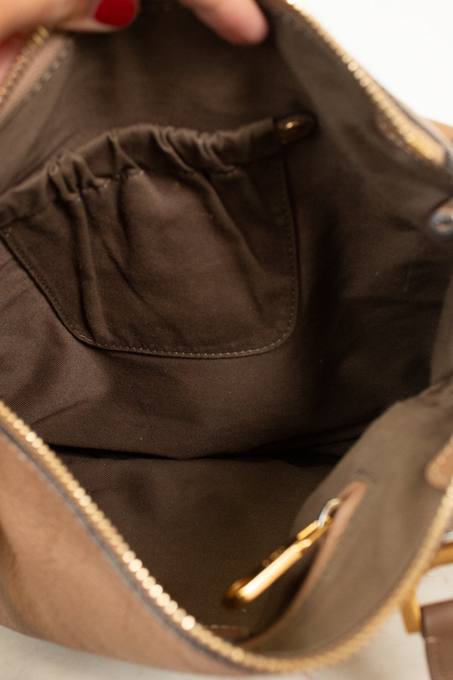 Close-up of the interior of a taupe-brown handbag, showing a dark brown fabric pocket with a gold-tone zipper pull and a gold zipper along the opening. A hand with red nail polish is holding the top edge.