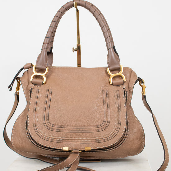 A structured taupe leather handbag with gold-tone hardware, dual top handles, and a detachable shoulder strap, displayed on a white marble block against a white background.
