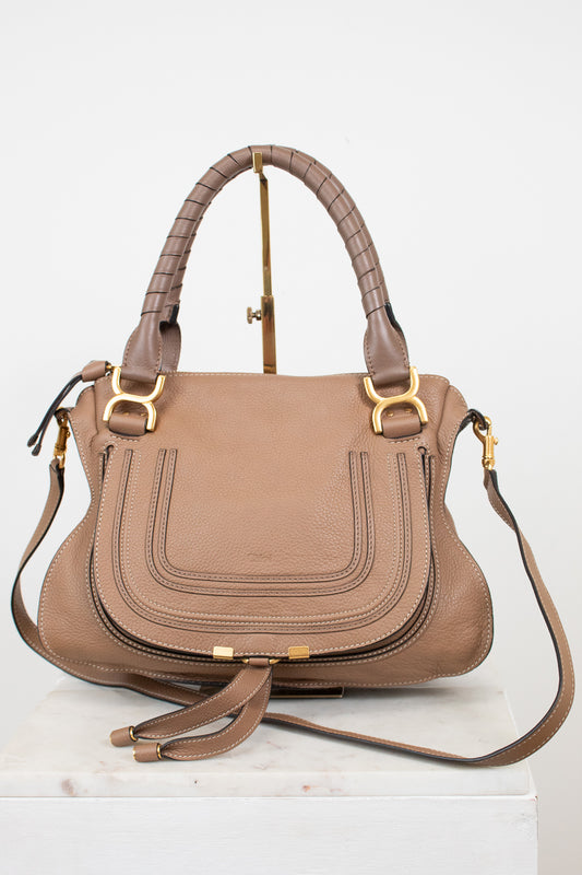 A structured taupe leather handbag with gold-tone hardware, dual top handles, and a detachable shoulder strap, displayed on a white marble block against a white background.
