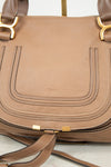 A close-up of a taupe brown pebbled leather handbag with gold-tone hardware and a matching leather strap resting in front.