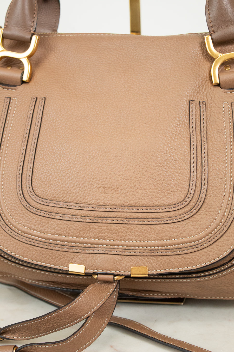 A close-up of a taupe brown pebbled leather handbag with gold-tone hardware and a matching leather strap resting in front.